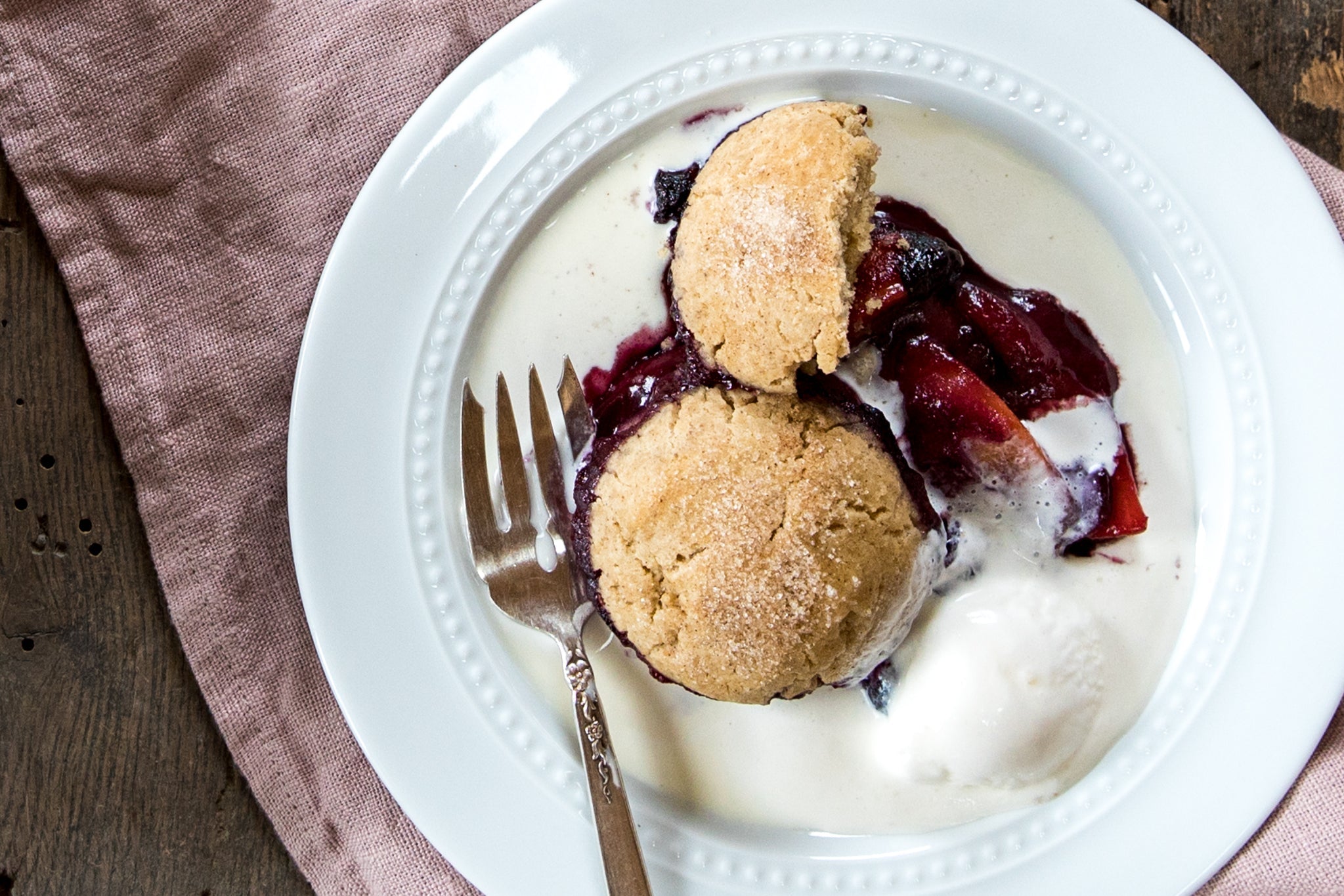 Einkorn peach and blueberry cobbler served with a scoop of vanilla ice cream on a white plate with a fork and pink napkin