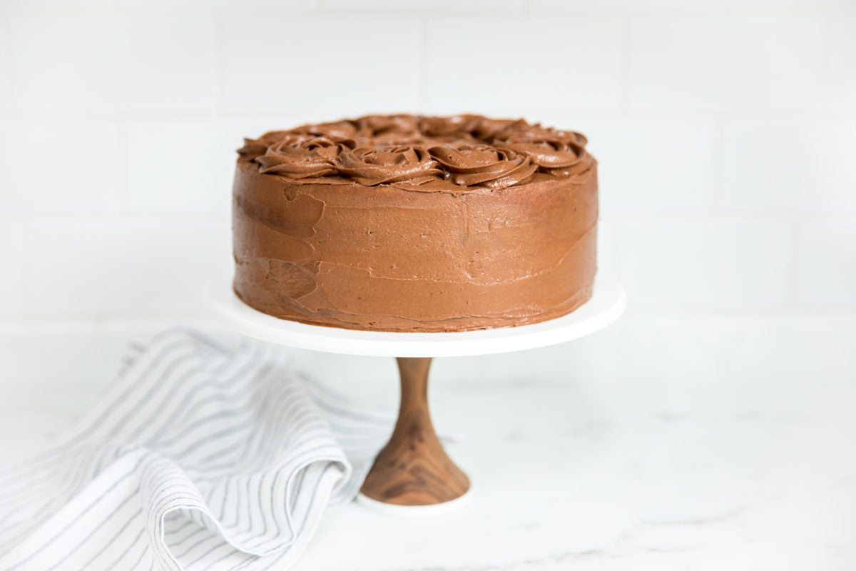 Side view of Einkorn yellow layer cake with chocolate buttercream, smooth sides and piped rosettes on a white pedestal with striped linen