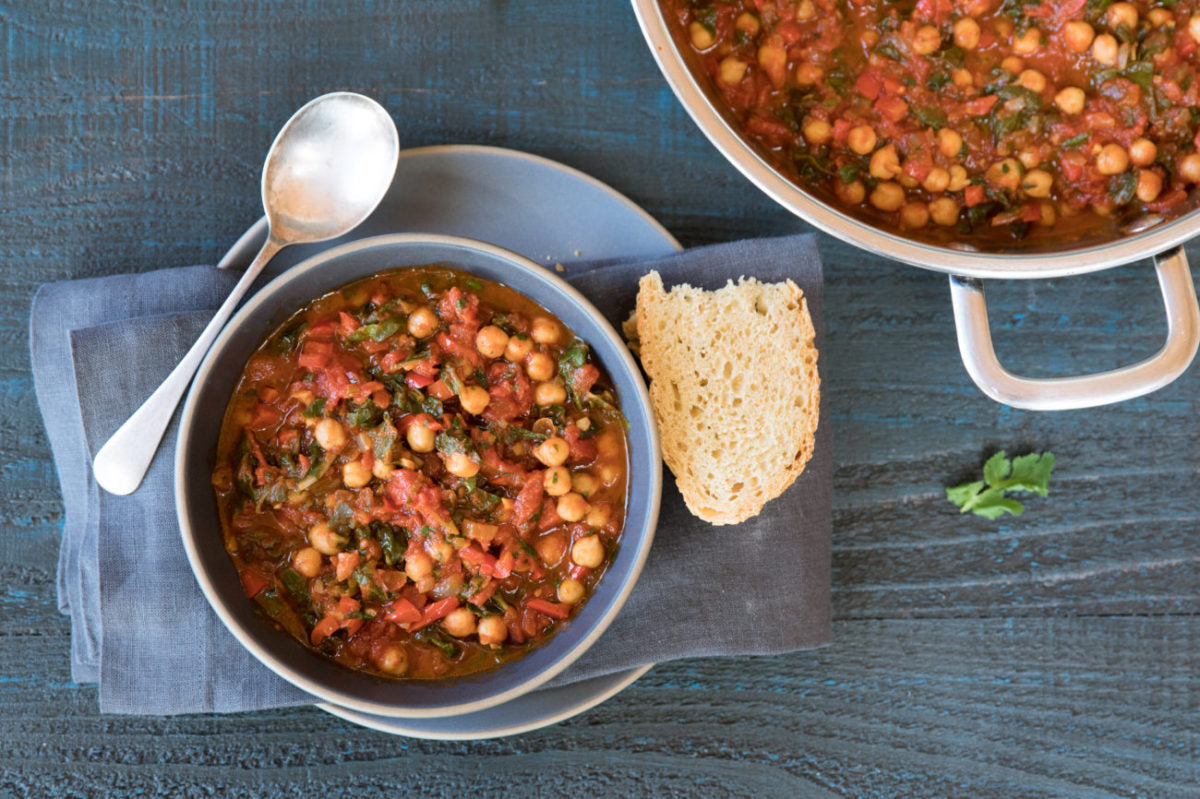 Bowl of chickpea and tomato stew on a blue napkin with a slice of bread and a pot of stew on a dark wooden table