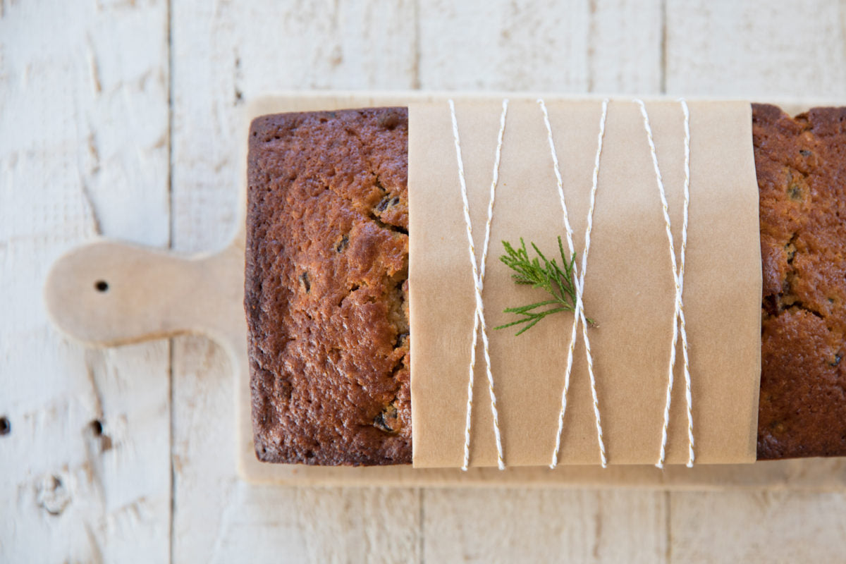 Overhead view of honey-sweetened rum fruit cake wrapped in paper and twine, garnished with an evergreen sprig on a board