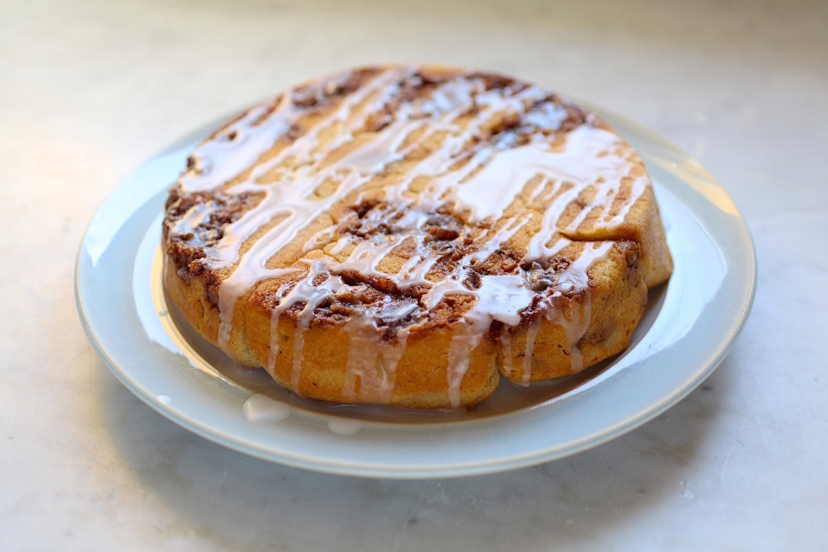 Three quarter view of a round Einkorn cinnamon roll on a white plate showing cinnamon swirls and icing drizzles on a marble surface