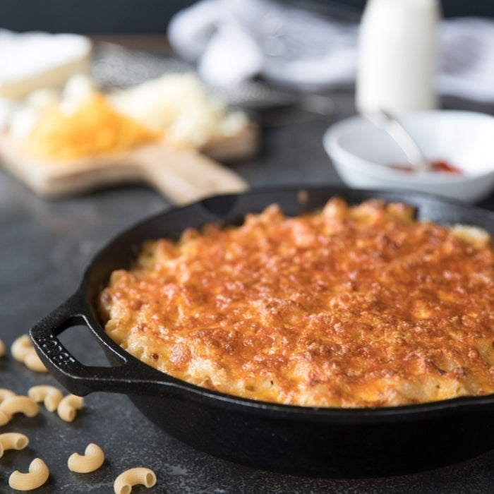 Baked mac and cheese in a cast-iron skillet with golden crust on a dark counter, elbow pasta and blurred cheese board