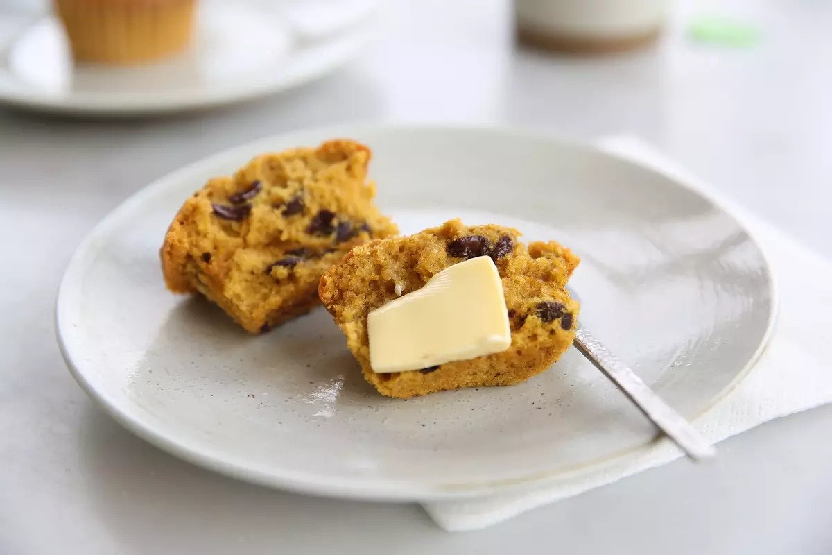 Two halves of an einkorn pumpkin chocolate chip muffin with a pat of melting butter on a white plate