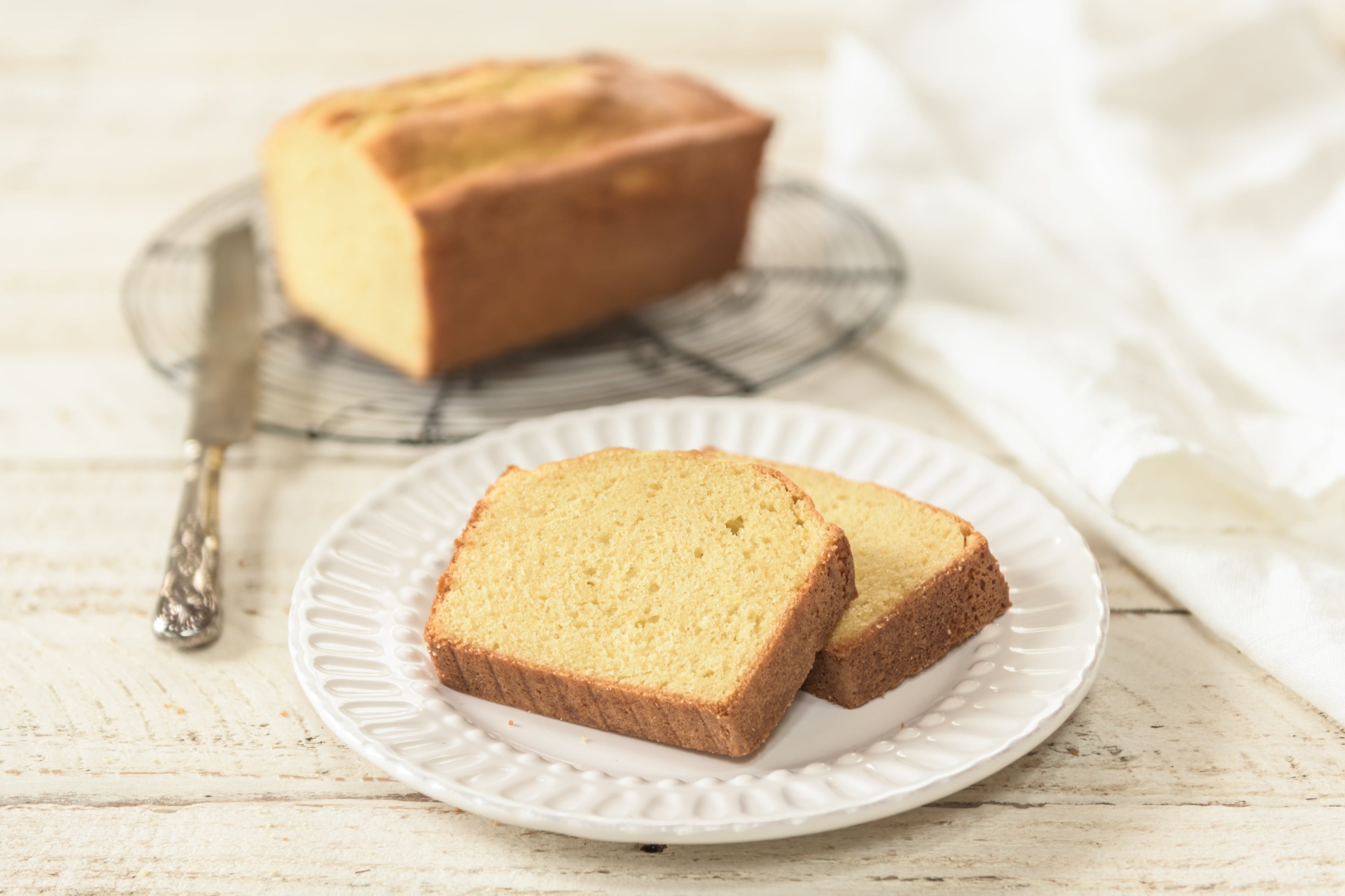 Two slices of einkorn pound cake on a white plate showing moist crumb and golden crust, loaf on a cooling rack behind