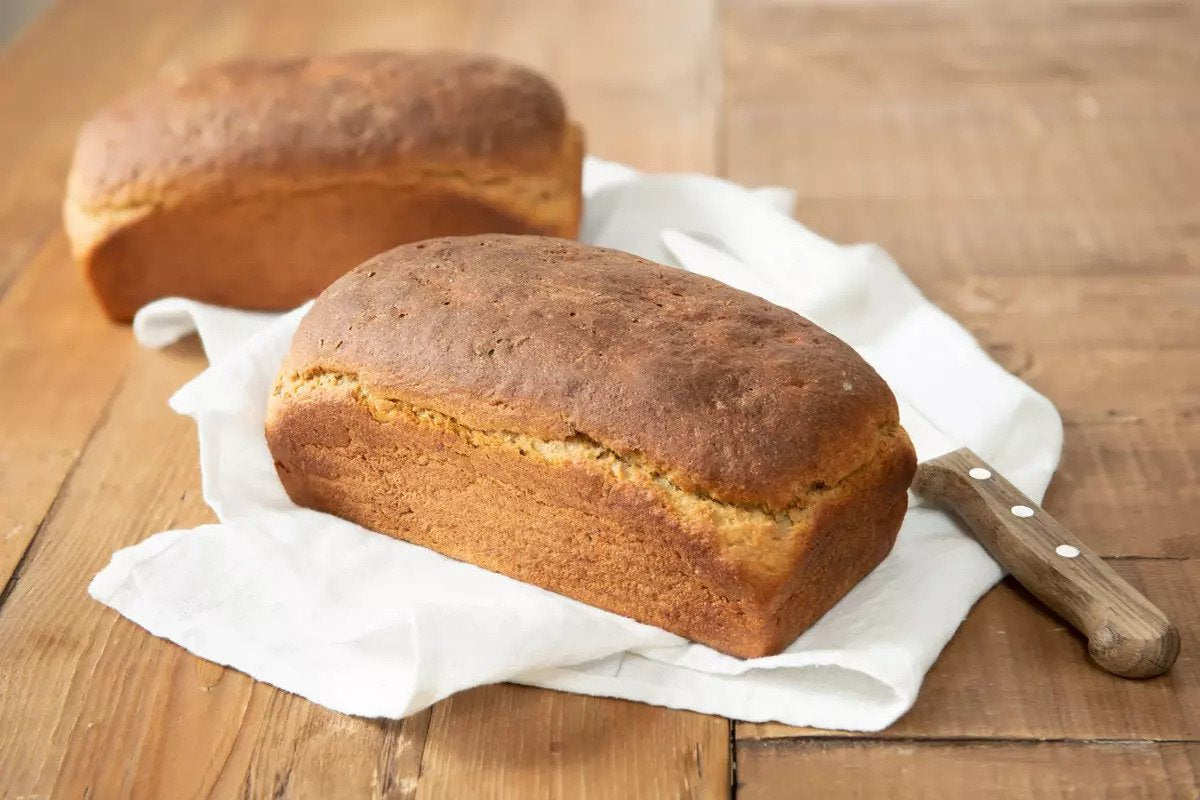 Front view of two Einkorn rye bread loaves showcasing golden-brown crusts on a white cloth with a wooden table and bread knife in the background