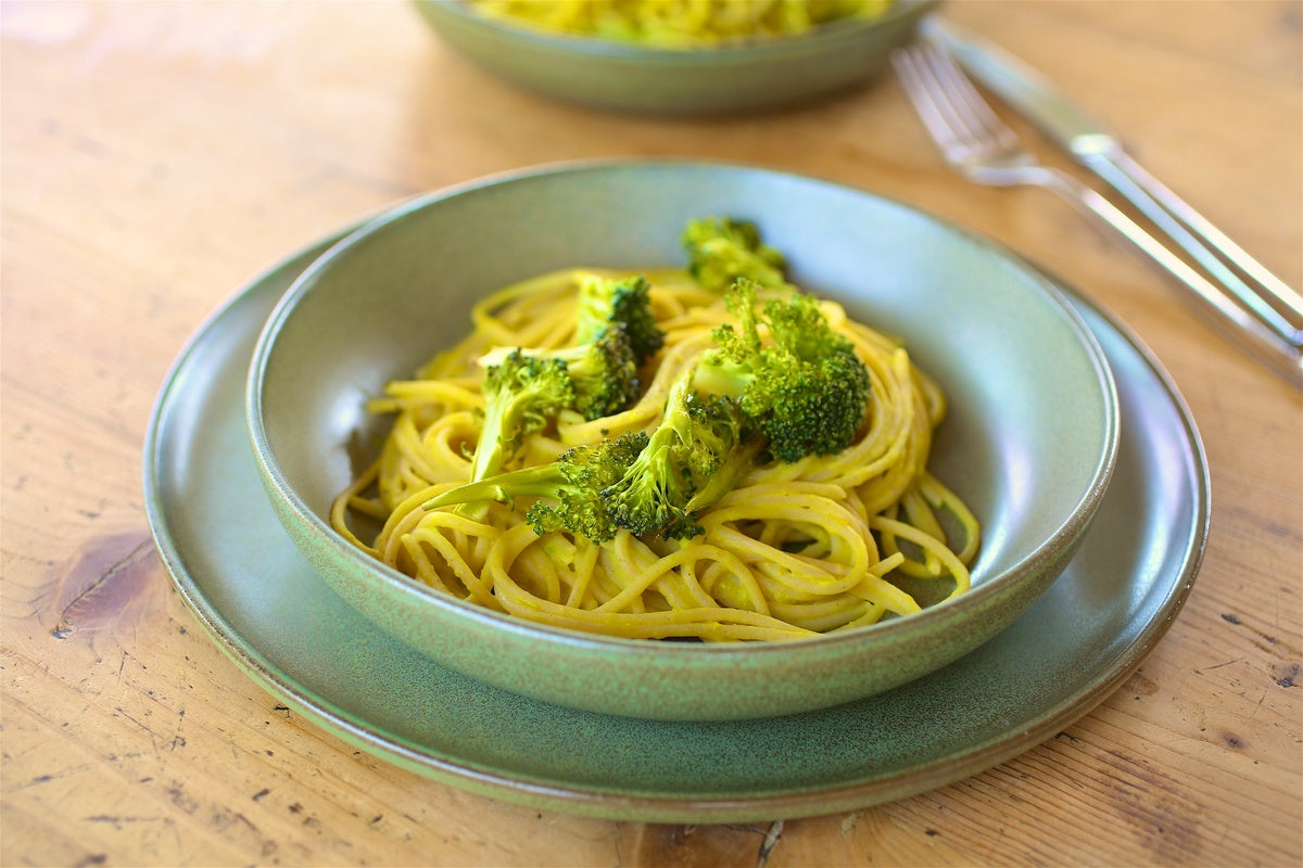 Bowl of spaghetti tossed in creamy chickpea sauce topped with roasted broccoli on a wooden table