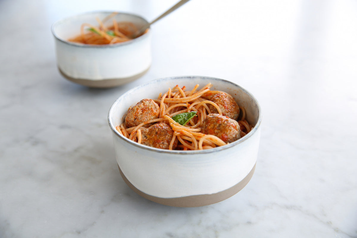 Front ceramic bowl of spaghetti and meatballs with basil on a marble countertop, second bowl blurred behind