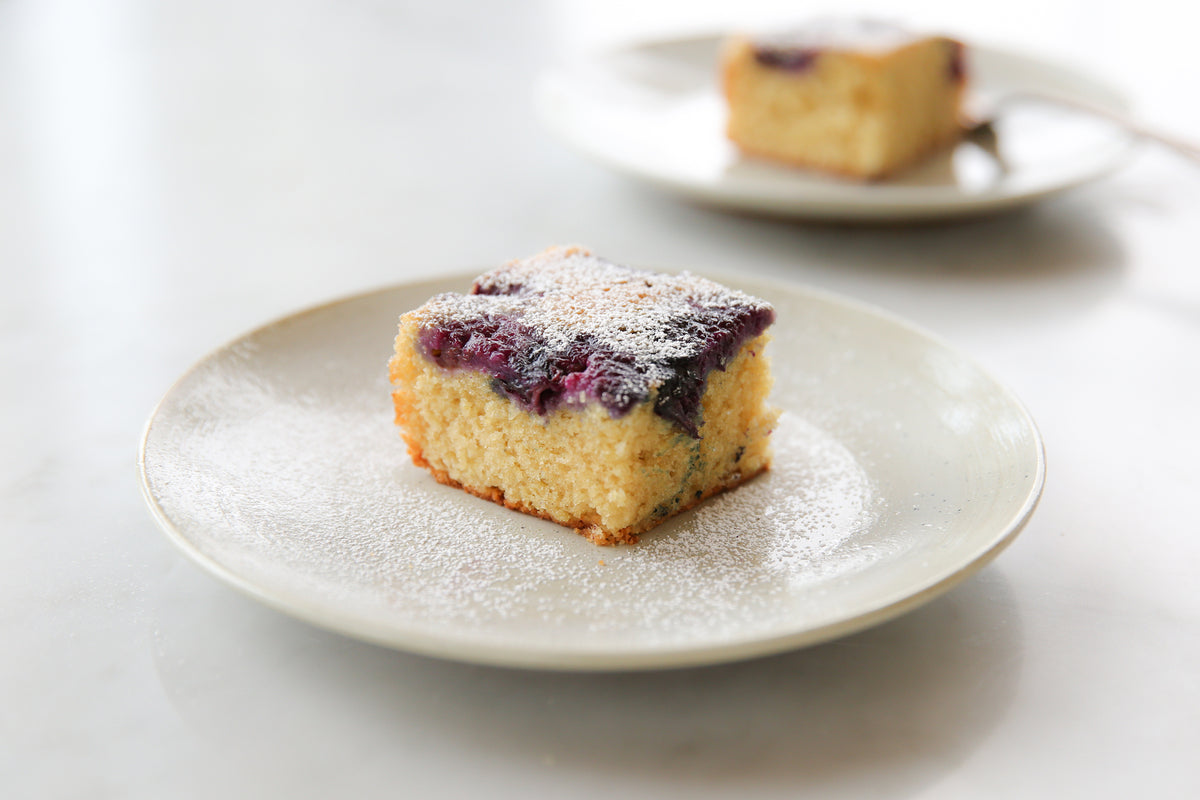 Close-up of a slice of Einkorn blueberry upside-down cake topped with blueberries and powdered sugar on a white plate