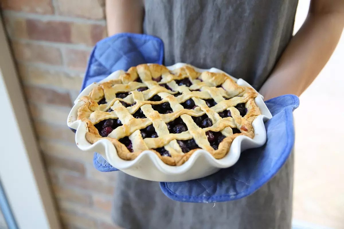 Person holding a lattice-top einkorn blueberry pie in a white fluted dish with blue oven mitts and a brick wall background