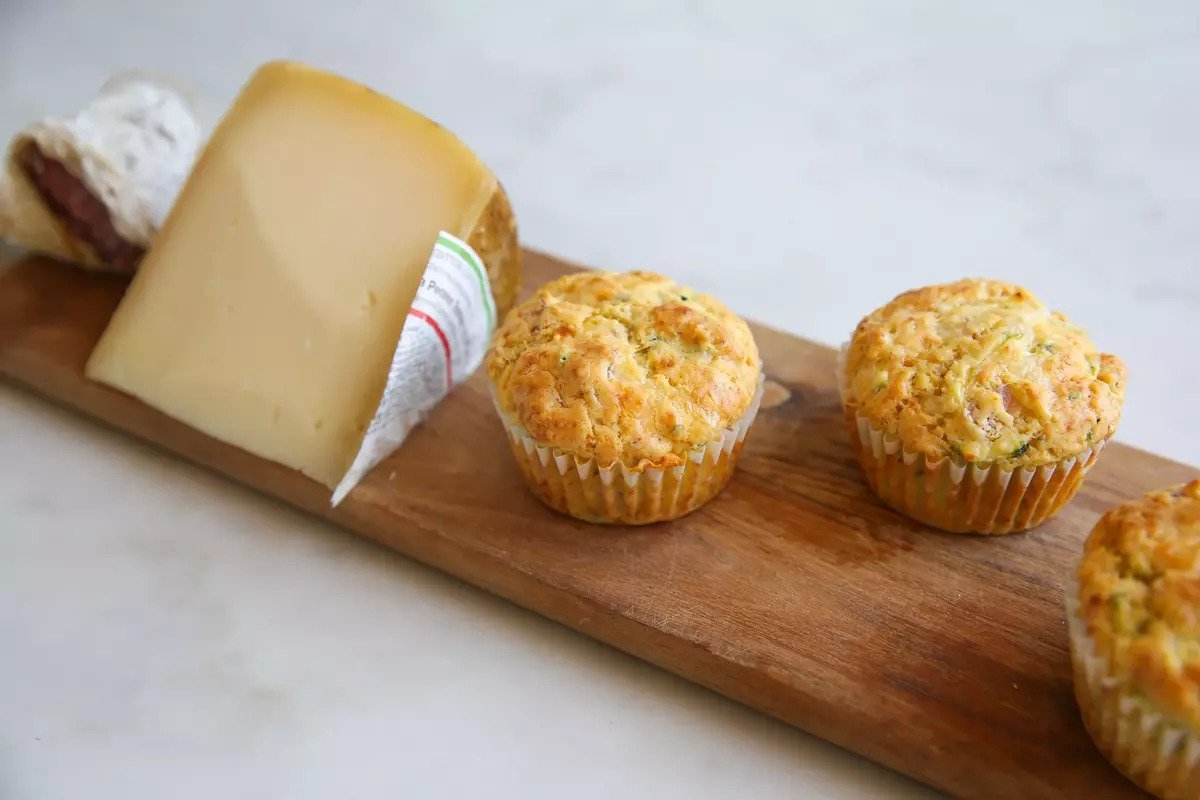 Three ham and cheese einkorn zucchini muffins on a wooden board with a wedge of cheese on a marble countertop