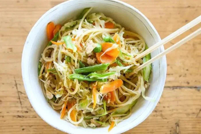 Bowl of teriyaki brown rice pasta stir-fry with cabbage, carrot ribbons and snap peas, chopsticks picking up noodles on a wooden table