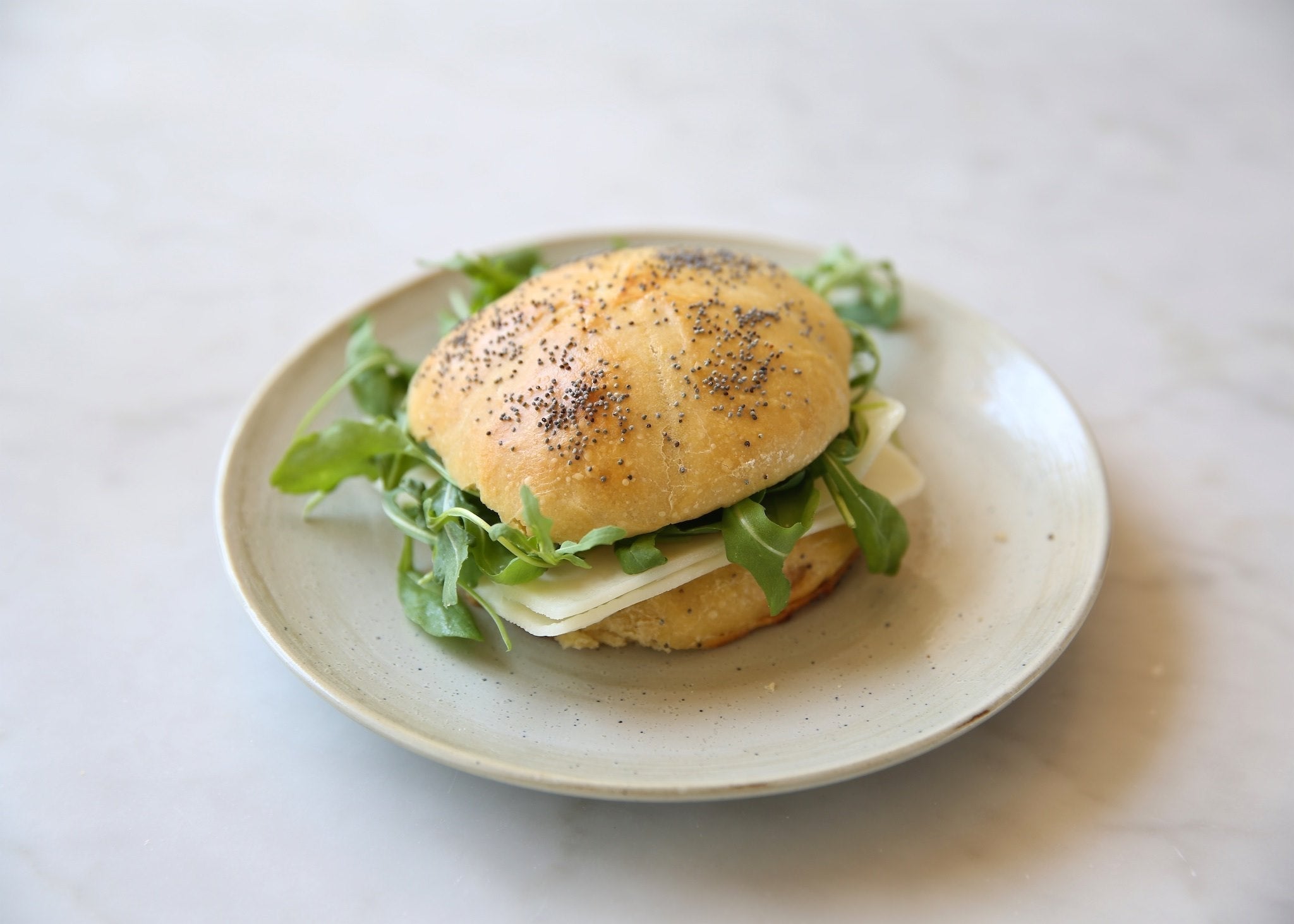 Angled view of Einkorn sourdough kaiser roll sandwich with poppy-seed bun, sliced cheese and arugula on a ceramic plate