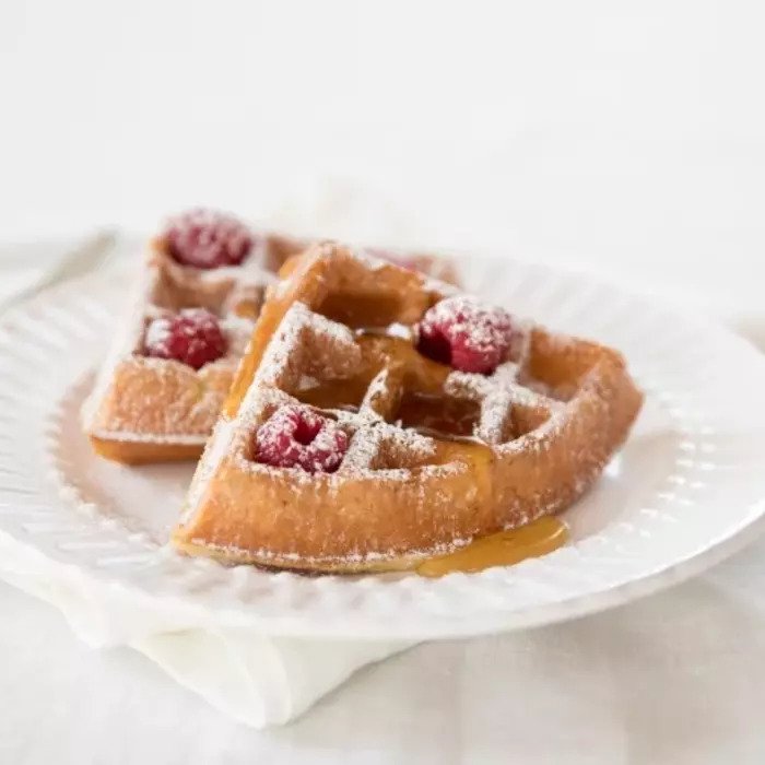 Two quartered Einkorn waffles dusted with powdered sugar, topped with raspberries and maple syrup on a white plate