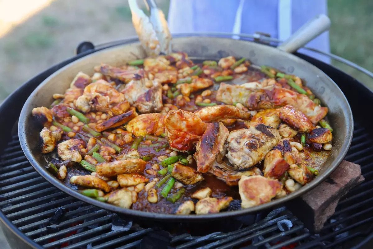 Paella cooking in a shallow pan on a charcoal grill with shrimp, chicken, green beans, and white beans