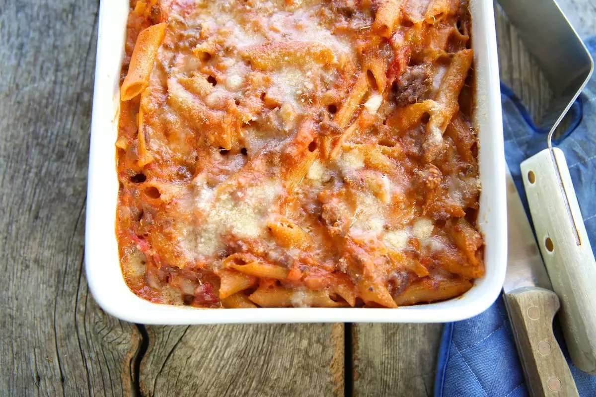 Baked penne Bolognese in white casserole dish topped with melted cheese, on a wooden table with blue napkin and spatula