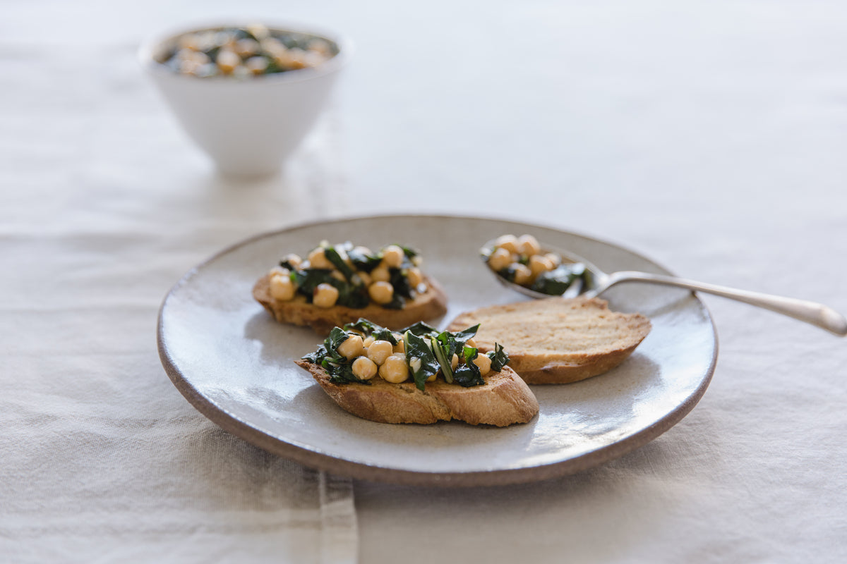 Garlicky chickpea and wilted greens on toasted bread slices on a ceramic plate with a spoon in soft natural light