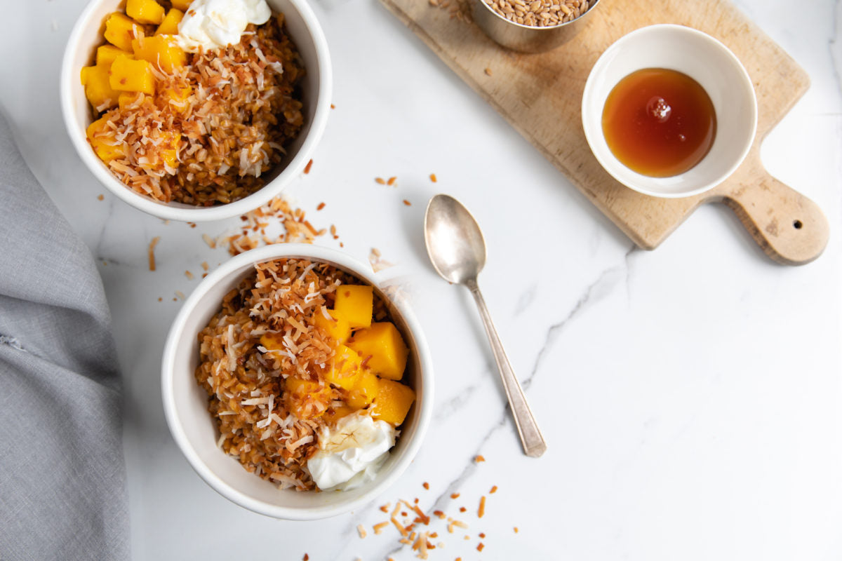 Two bowls of wheat berry breakfast topped with mango cubes, toasted coconut flakes, and yogurt on a marble countertop