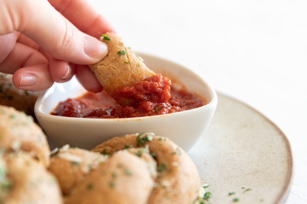 Hand dipping a whole-grain einkorn garlic knot into a bowl of marinara sauce on a plate