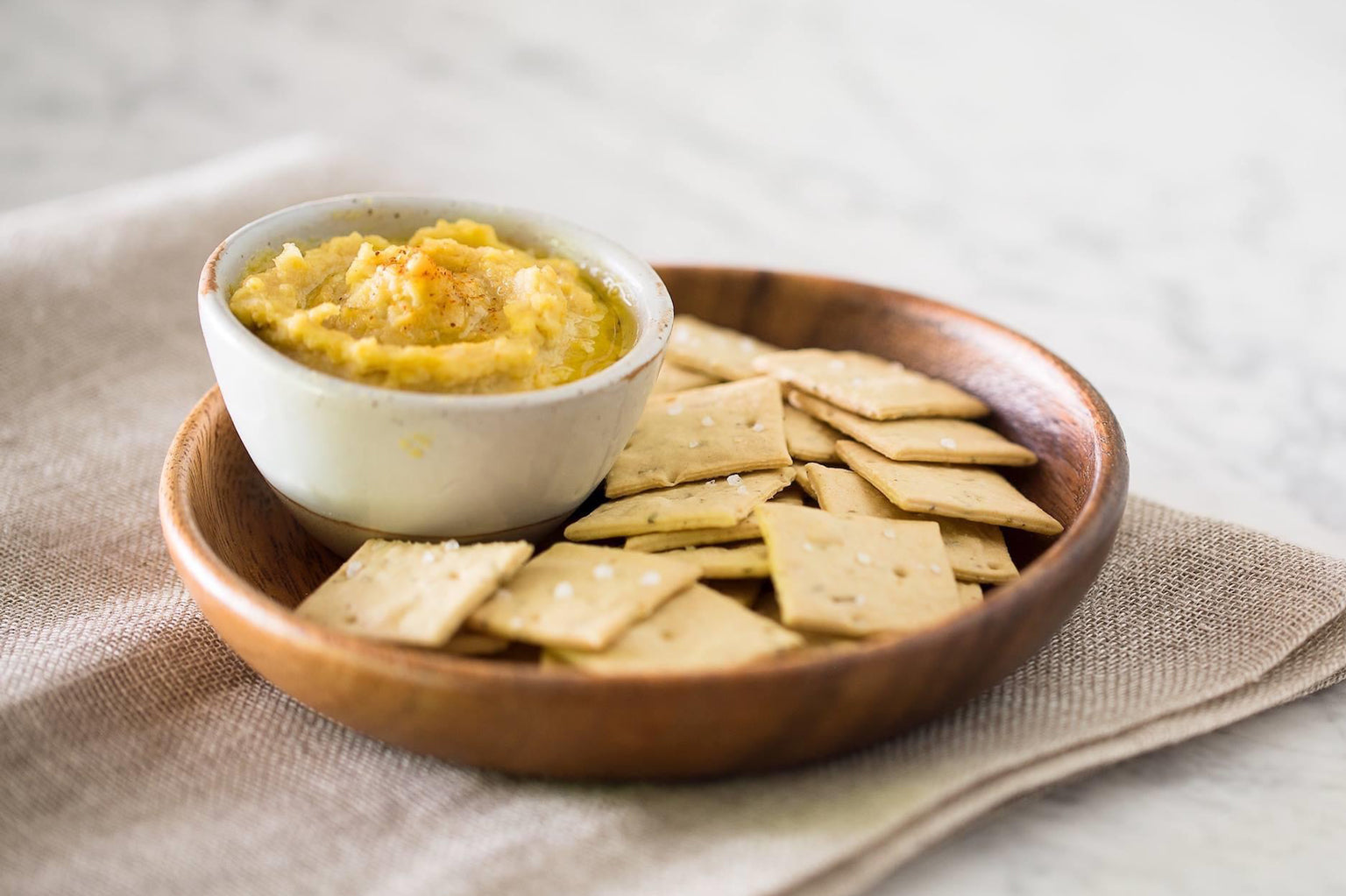 Bowl of hummus drizzled with extra-virgin olive oil served with sea-salt crackers on a wooden plate