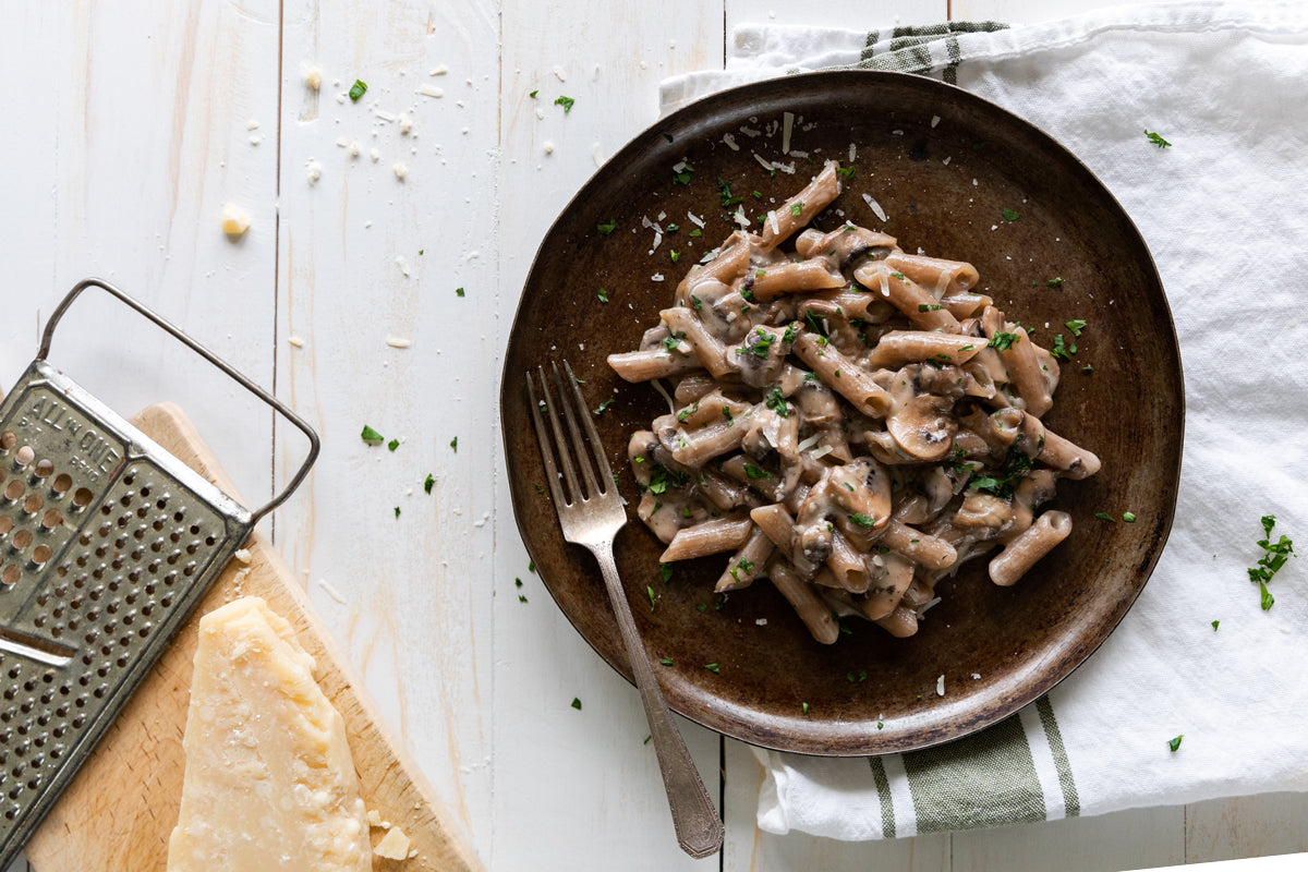 Creamy grain-free mushroom penne served on a brown plate with a fork on a white wooden table, sprinkled with grated cheese and parsley