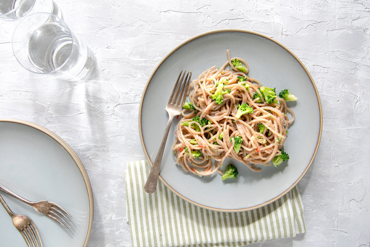 Plate of gluten-free Jovial cassava spaghetti with garlic, chili flakes, and broccoli on a light gray plate with a fork and striped napkin