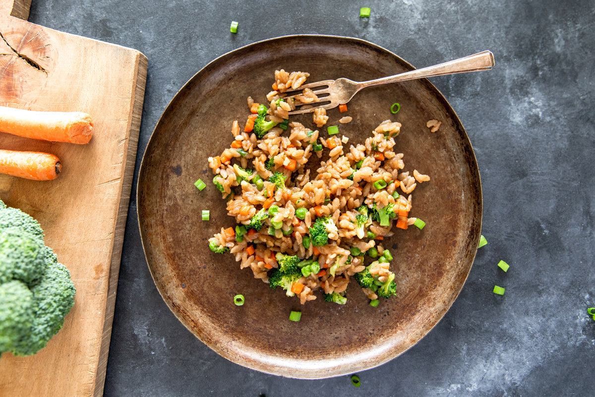Gluten free cassava orzo “fried rice” with broccoli, diced carrots and scallions on a rustic brown plate over a dark stone surface