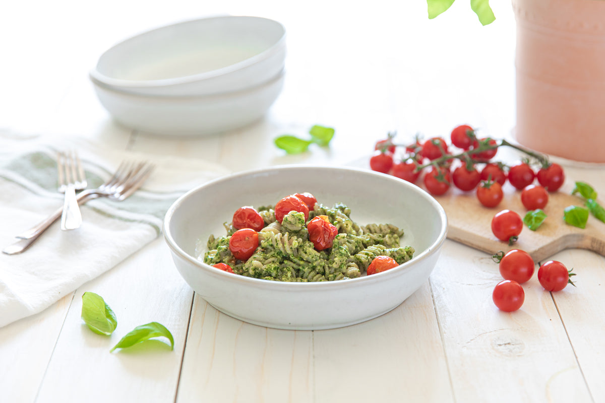 Bowl of cassava fusilli tossed in cashew pesto with roasted cherry tomatoes on a bright white table with basil and forks