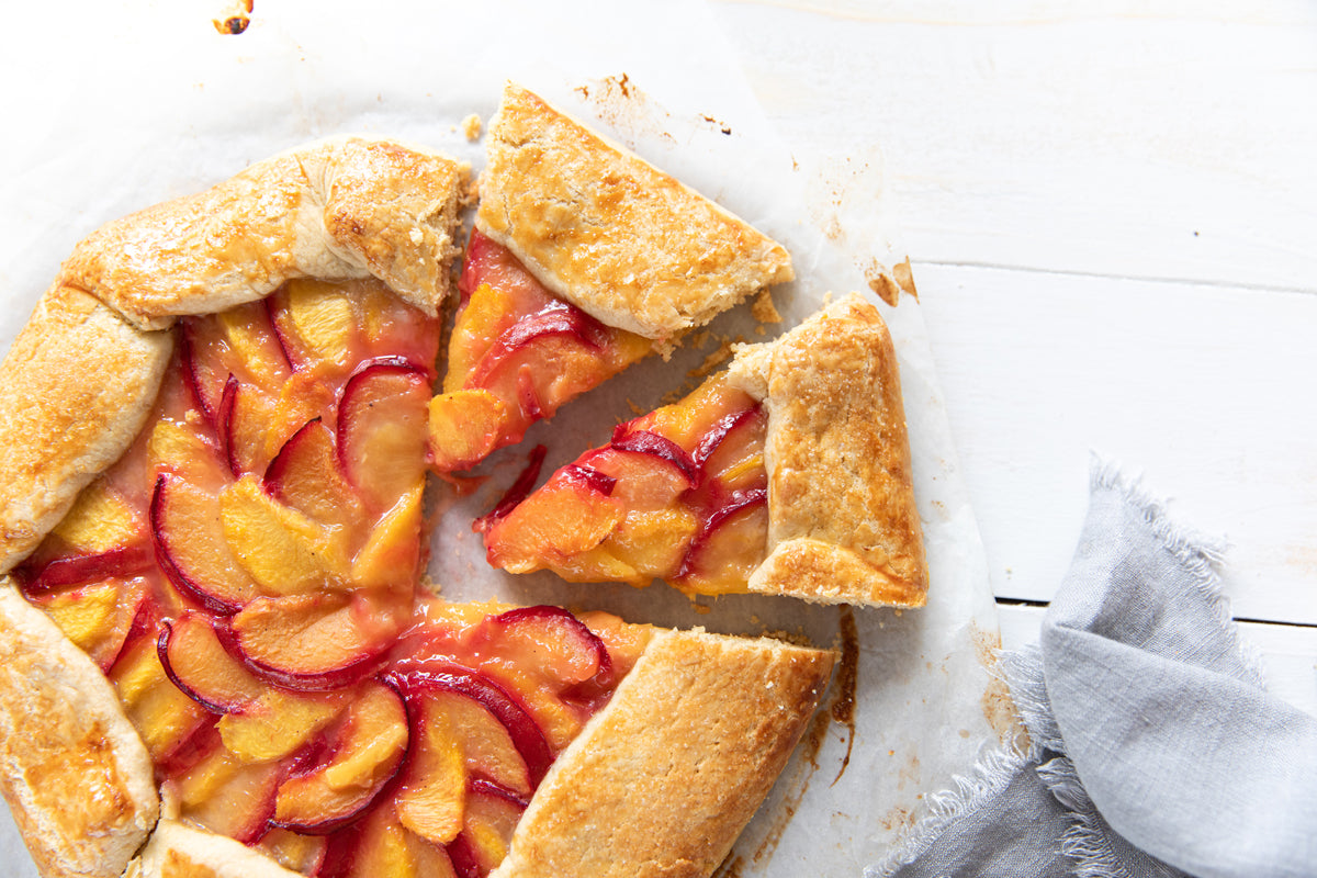 Einkorn summer fruit galette with sliced peaches and plums, one wedge removed on parchment on white wood with gray linen