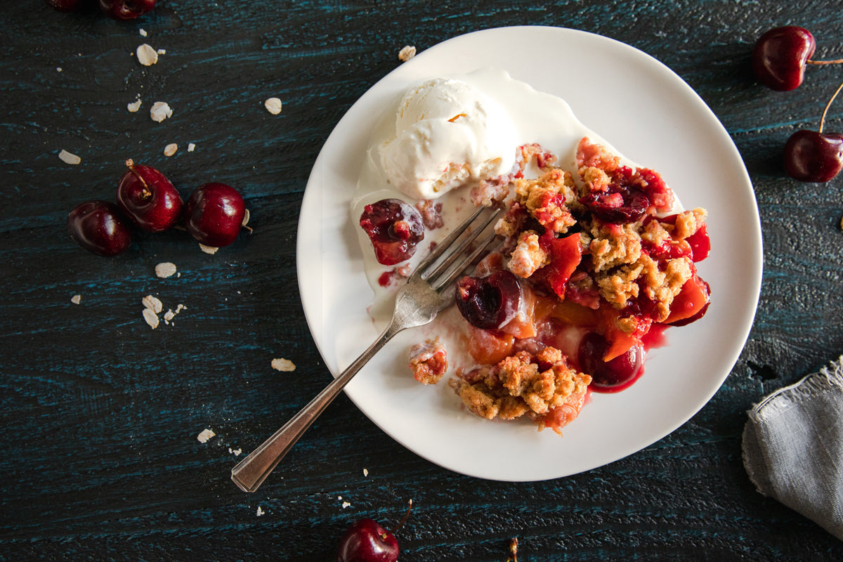 Plate of Einkorn fruit crumble with peaches and cherries and a scoop of vanilla ice cream on a dark wooden table