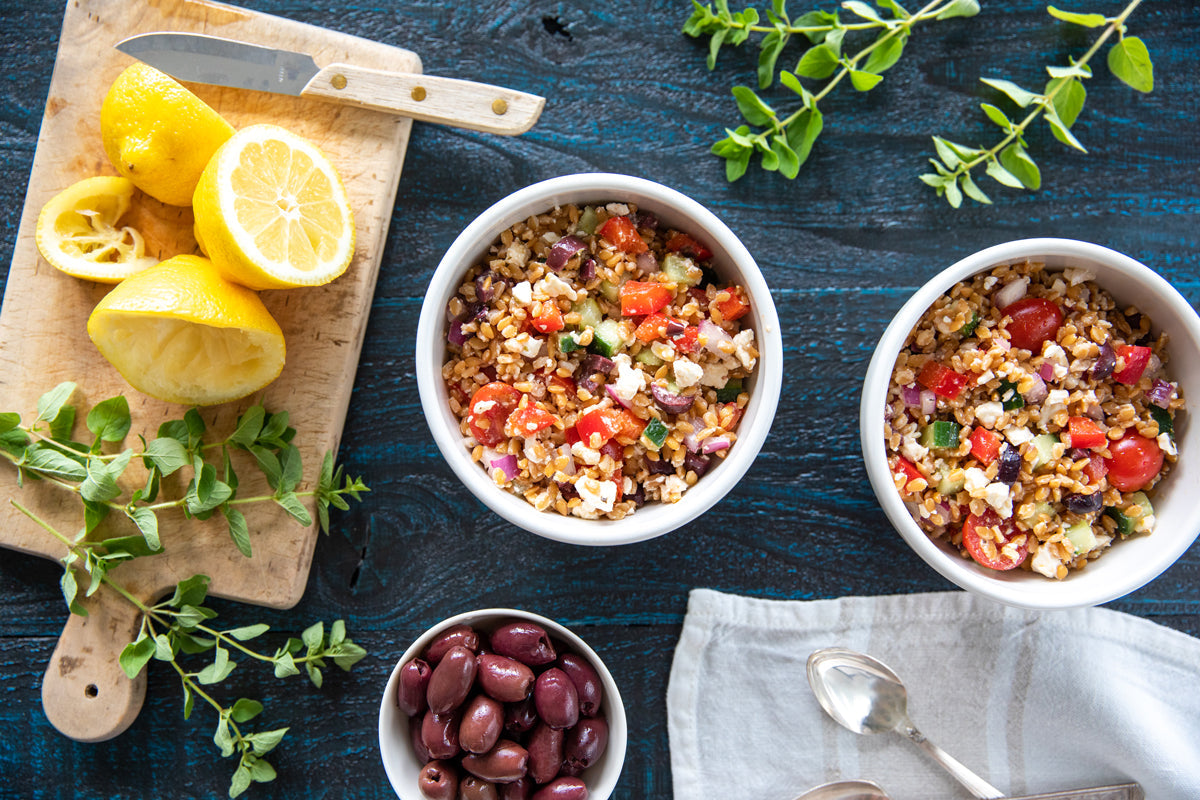 Top-down view of two bowls of Greek Einkorn wheat berry salad with tomatoes, cucumber, red onion and feta on a dark blue wooden table