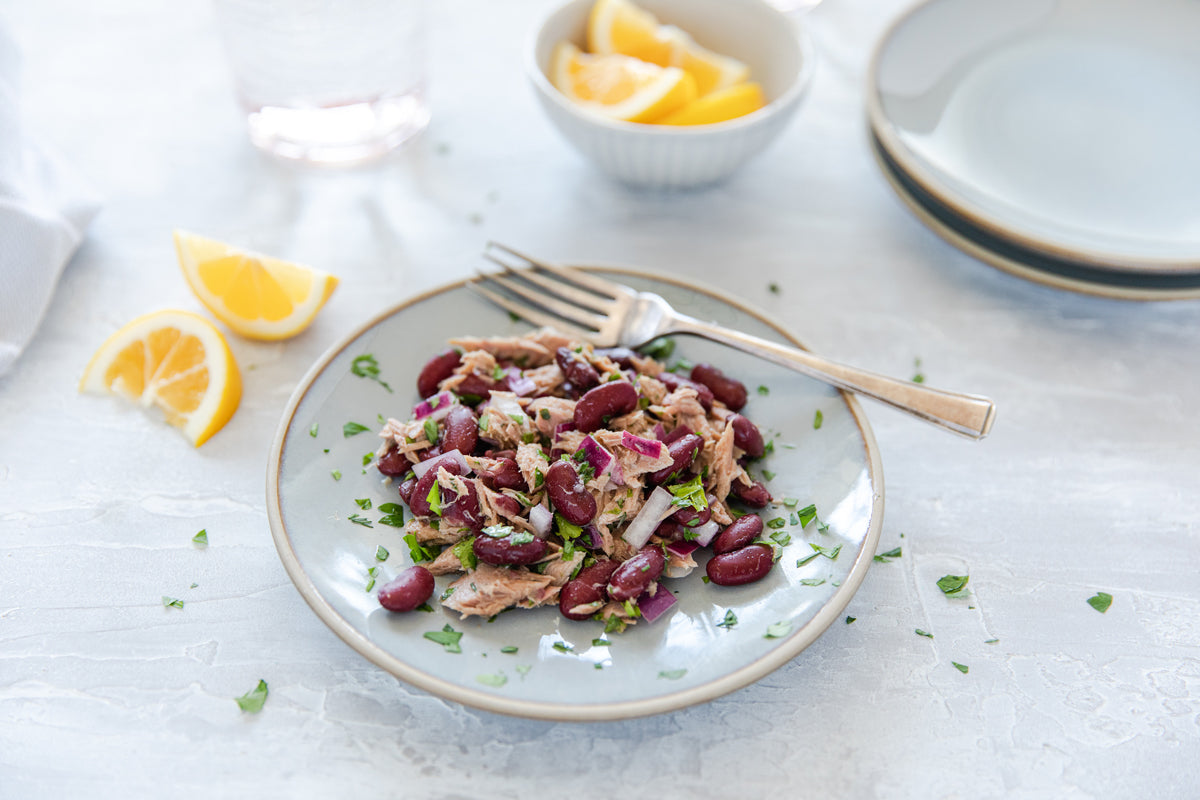 Tuna and kidney bean salad with red onion and parsley on a gray plate, fork and lemon wedges on a light table