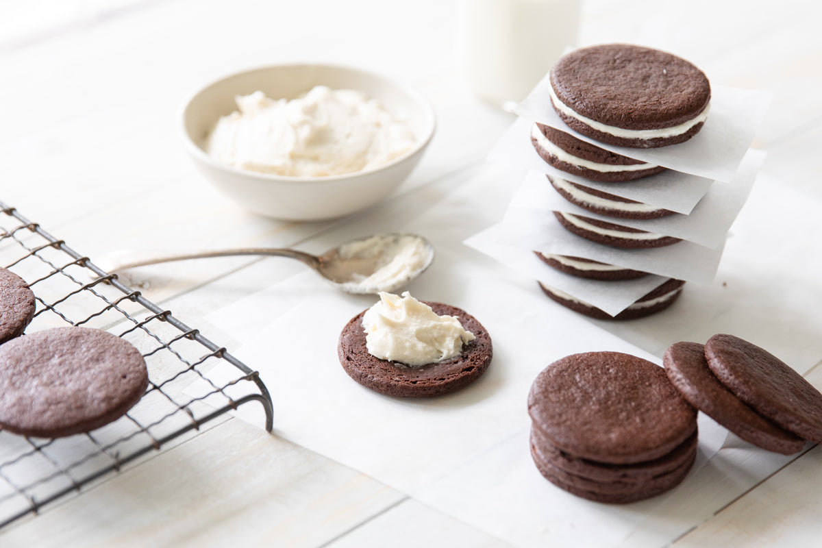 Einkorn chocolate sandwich cookies on a white table, one cookie spread with frosting, stack of cookies and bowl of cream