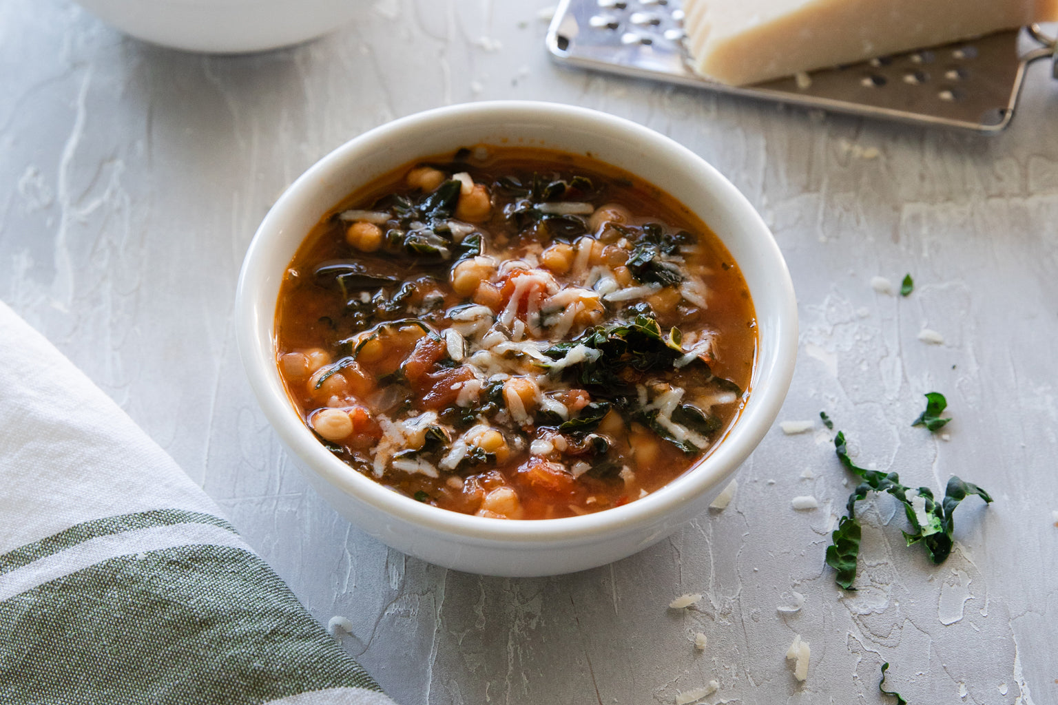 Bowl of kale, chickpea, and tomato soup topped with grated cheese on a gray surface with a towel and cheese grater