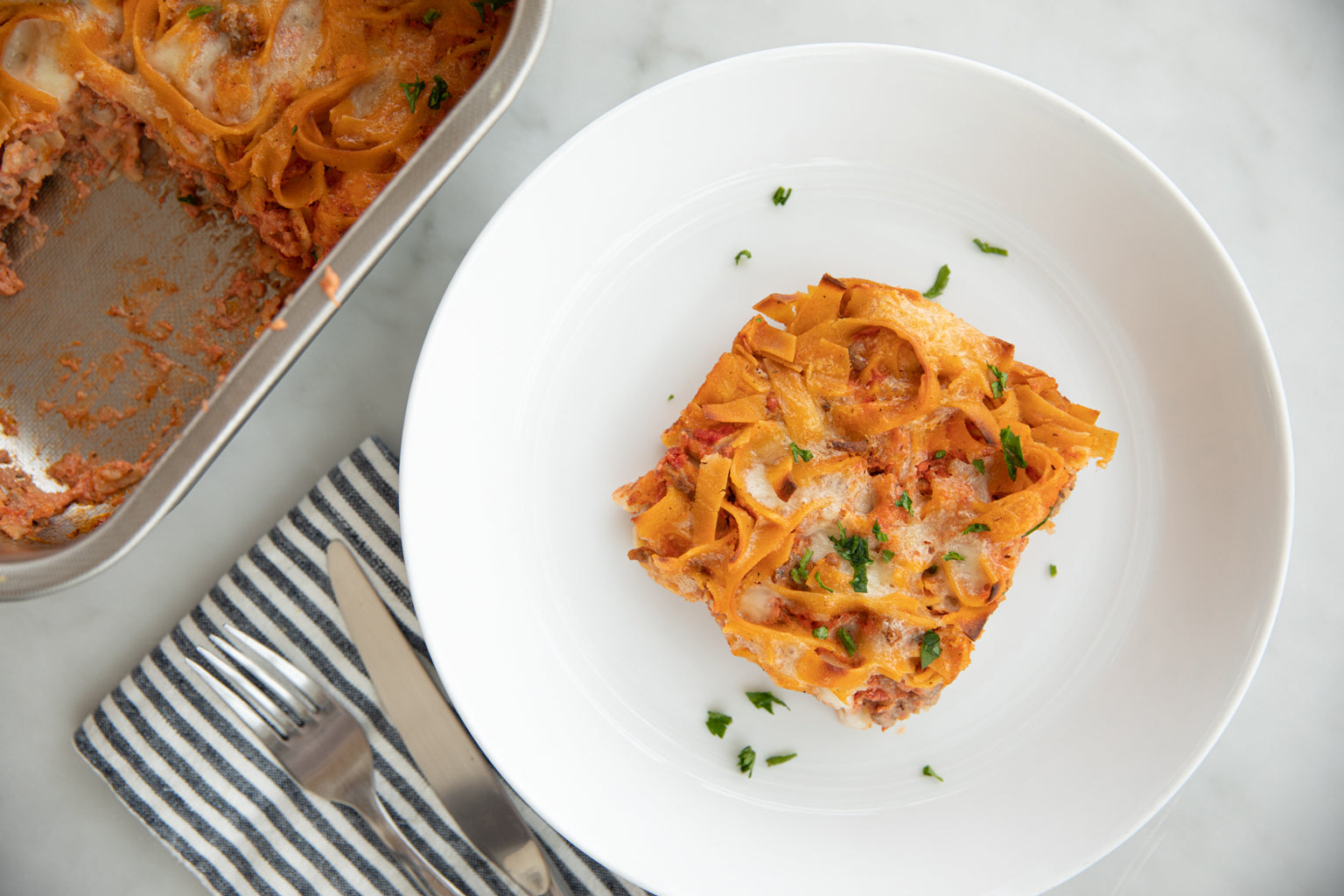 Slice of gluten-free noodle casserole served on a white plate next to a metal baking pan and striped napkin