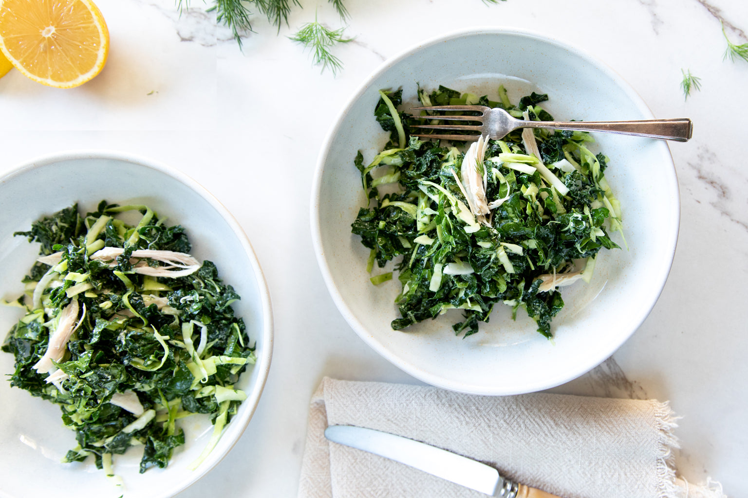 Two bowls of winter kale salad with shredded chicken and shaved fennel on a marble tabletop, fork in one bowl