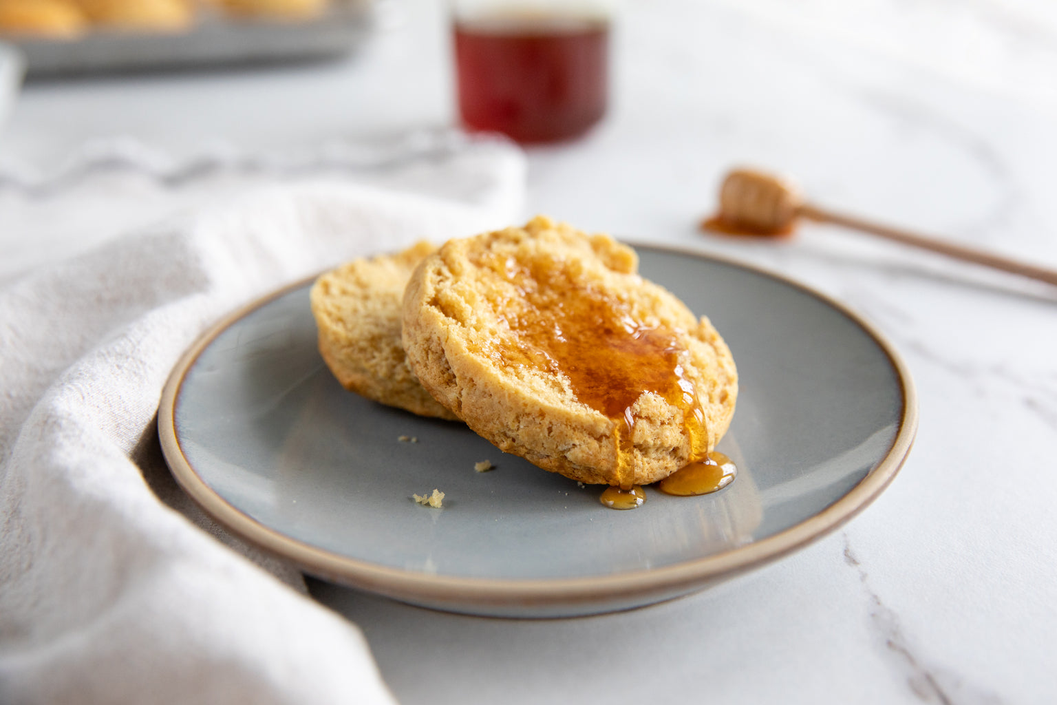Two honey-drizzled einkorn biscuits on a gray plate with a jar of honey and honey dipper blurred in the background