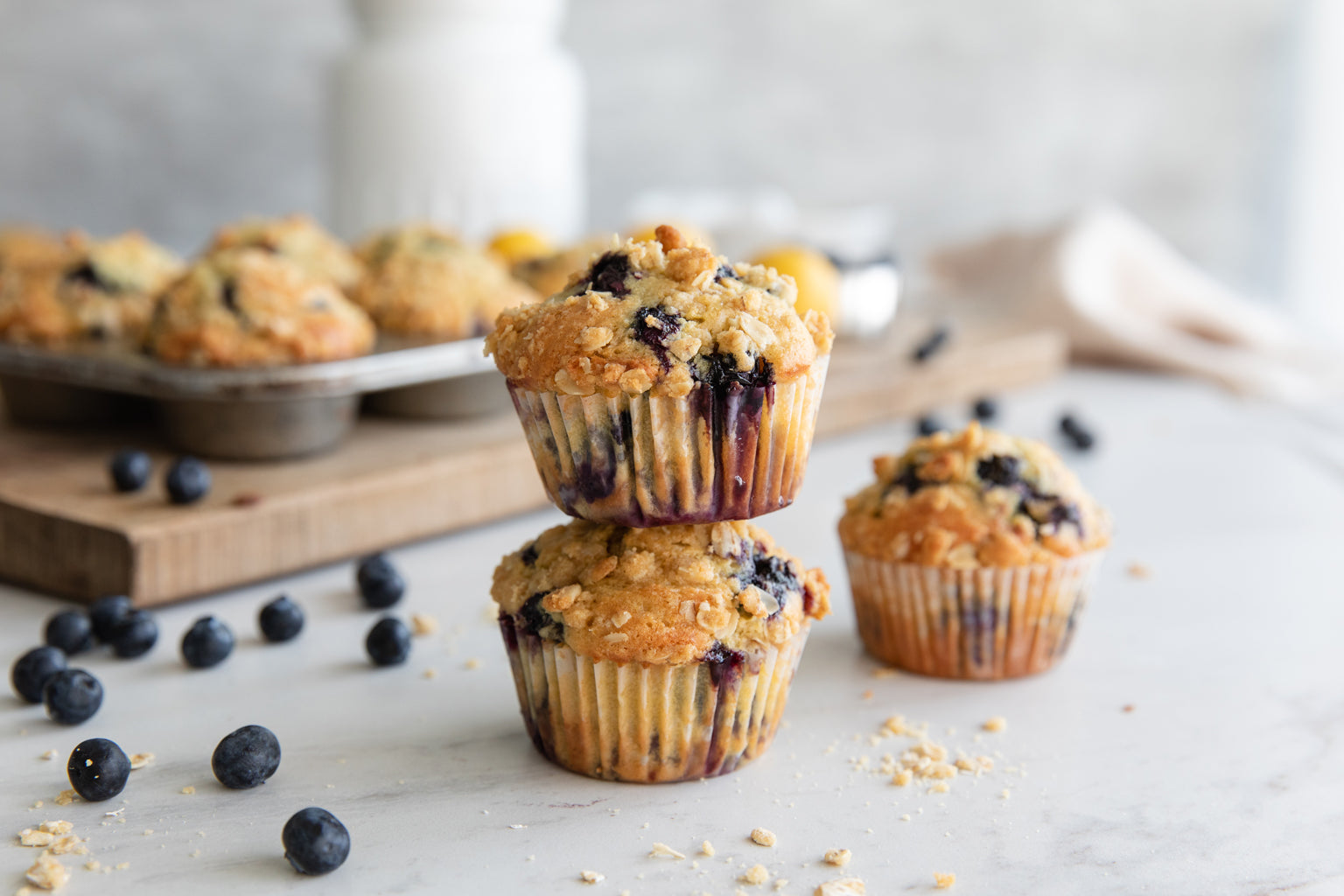 Einkorn blueberry streusel muffins stacked on a marble surface with scattered blueberries and a blurred muffin tin in the background