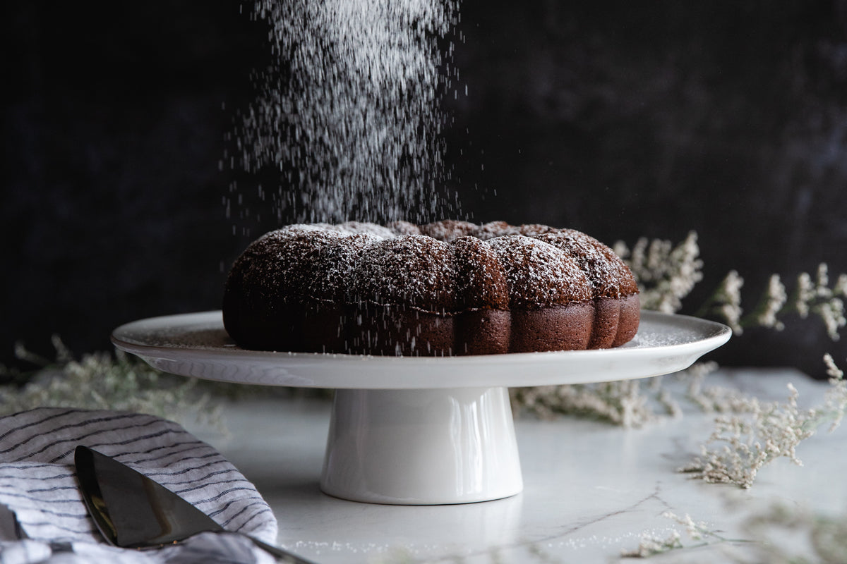 Chocolate olive oil Bundt cake being dusted with powdered sugar on a white ceramic cake stand against a dark backdrop