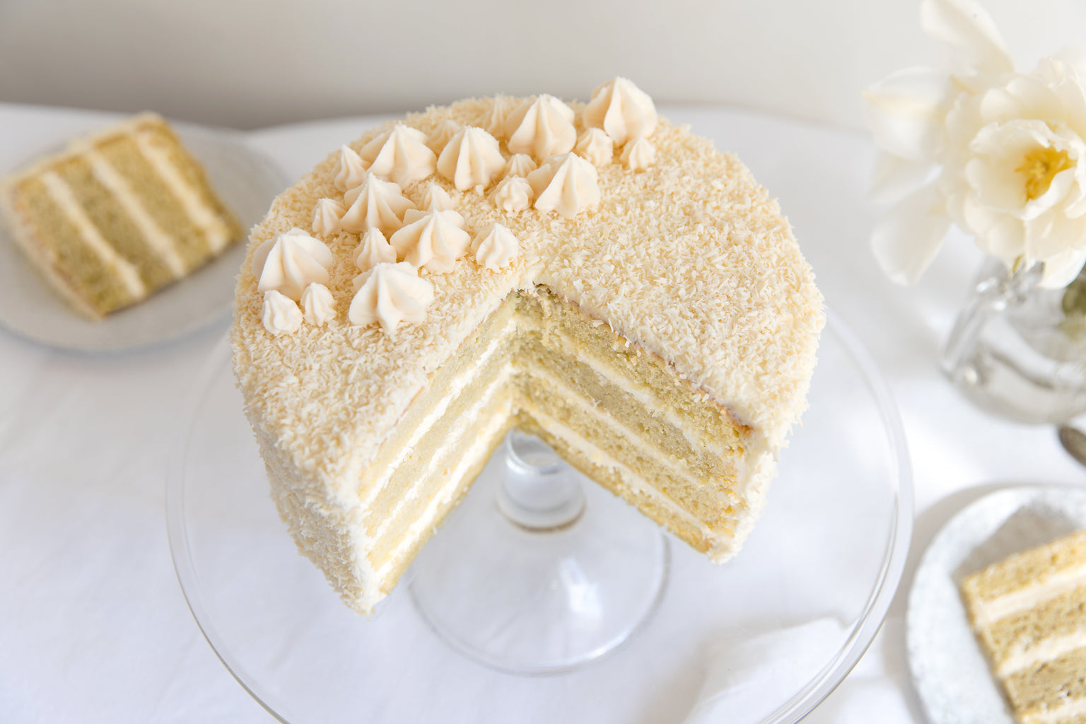 Angled overhead view of an Einkorn coconut layer cake with four layers, coconut frosting and piped dollops on a glass stand