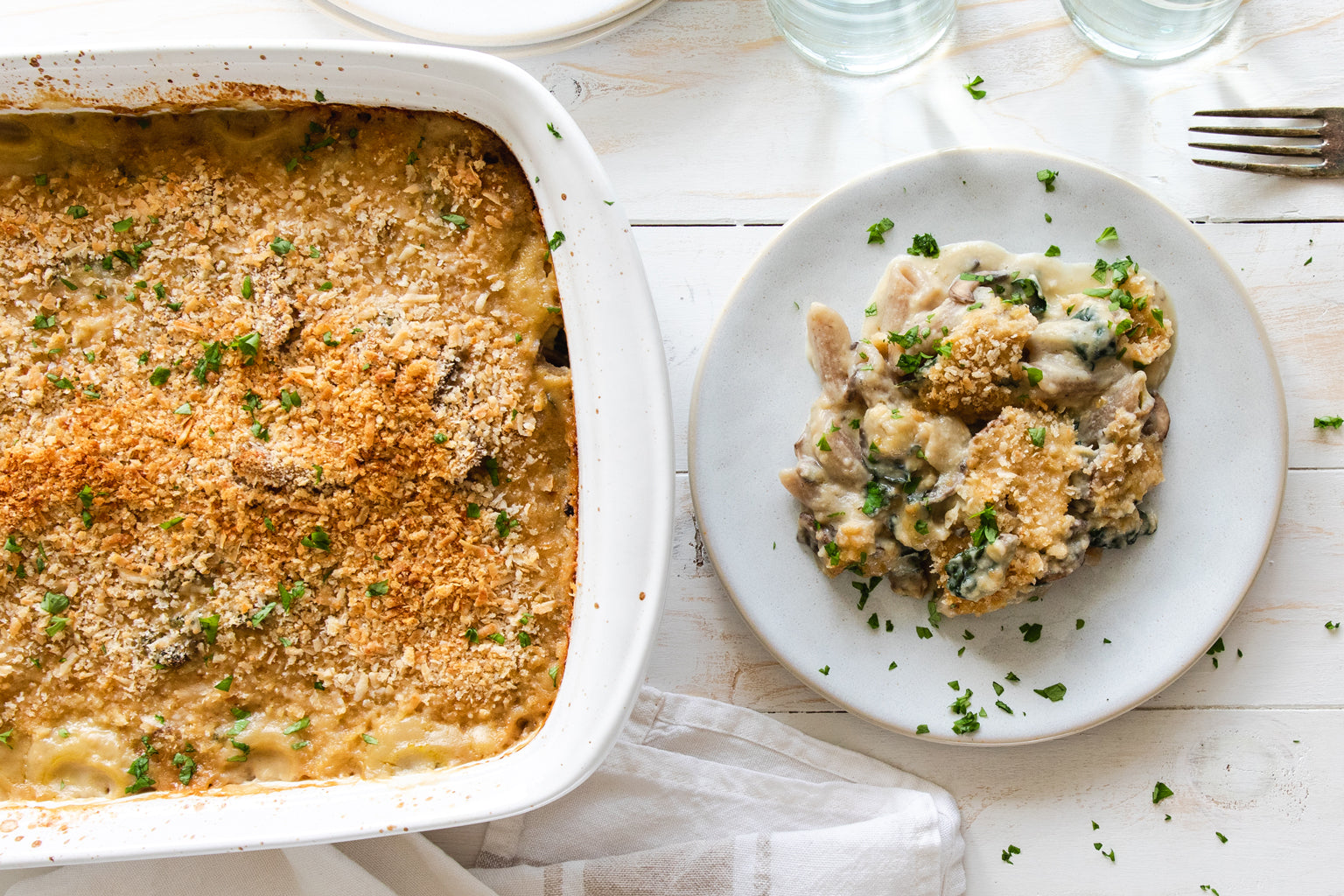 Baked penne in creamy cauliflower sauce with kale and mushrooms, plated beside a breadcrumb-topped casserole dish
