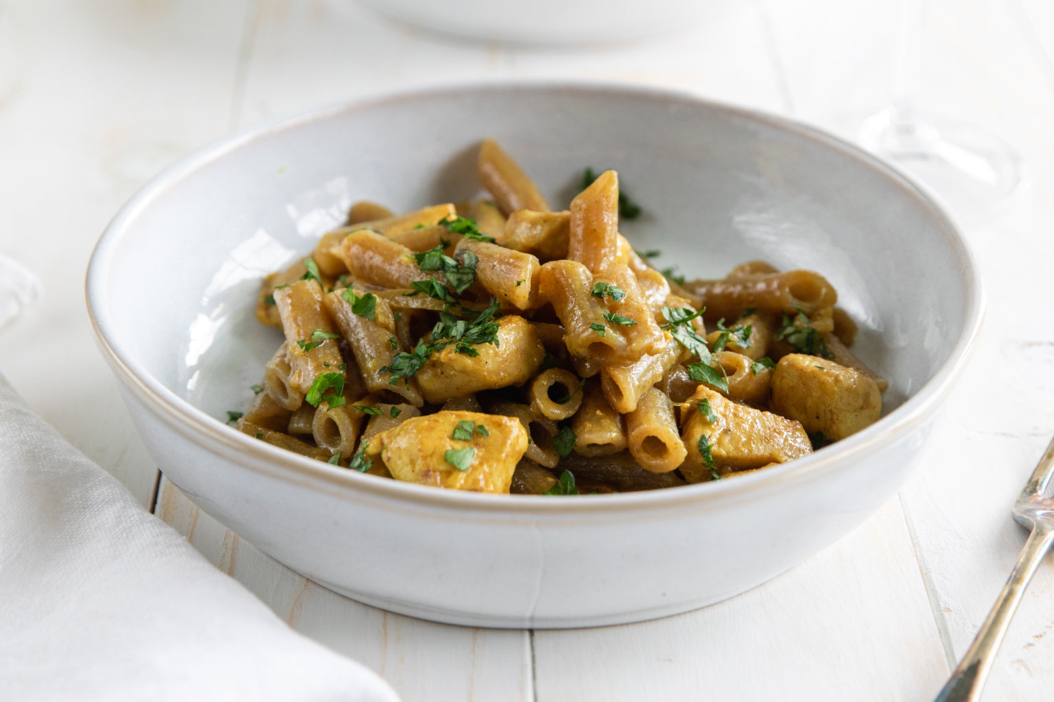 Bowl of coconut curry chicken with grain-free cassava penne, garnished with chopped parsley on a white table