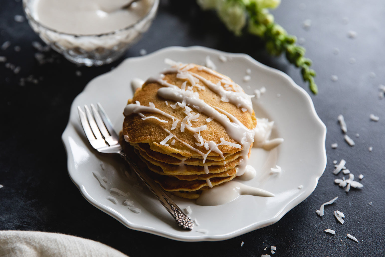Stack of einkorn coconut pancakes drizzled with coconut cream and sprinkled with shredded coconut on a white plate with fork