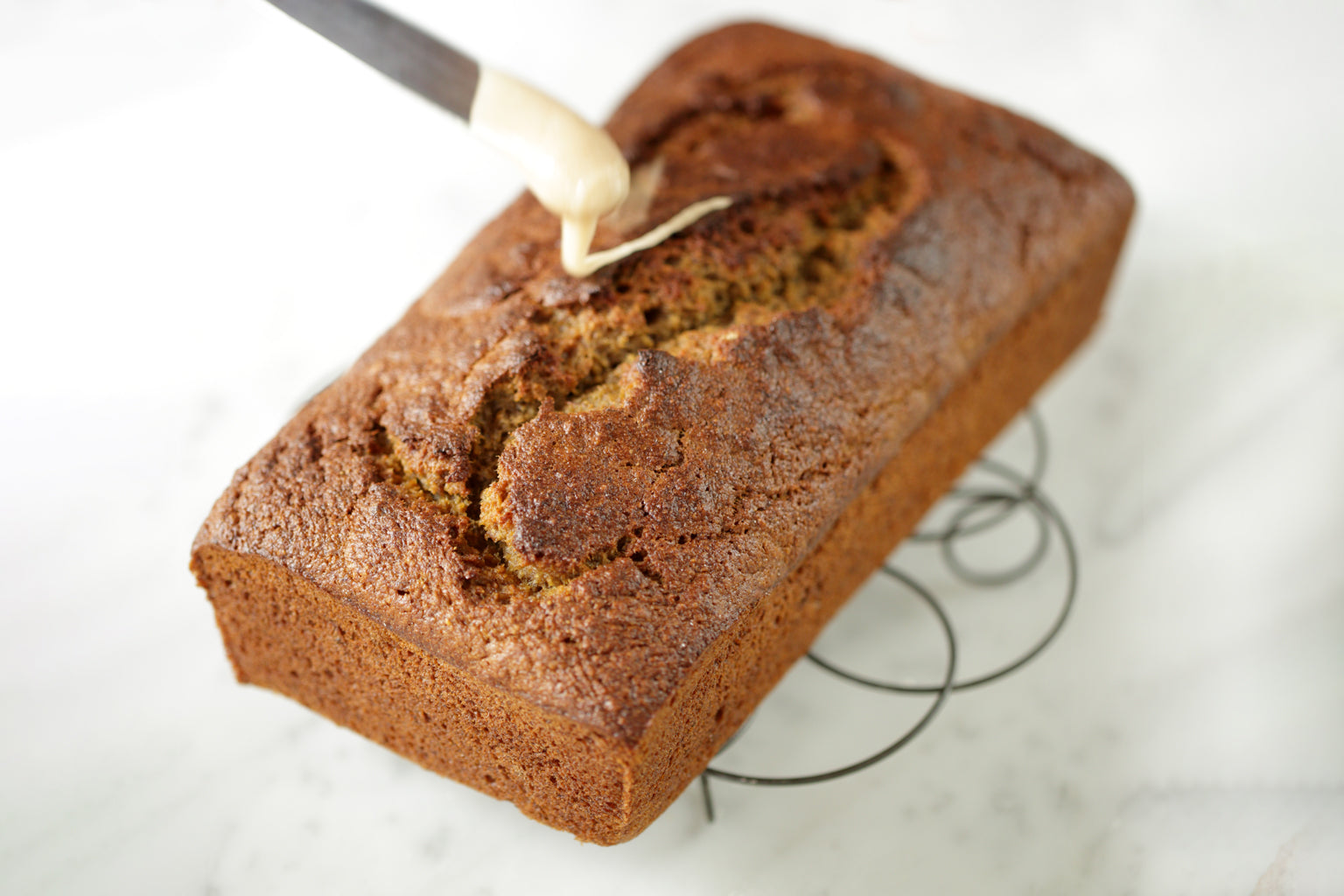 Angled top-down view of Einkorn banana bread with cracked golden crust and glaze being drizzled on a wire cooling rack