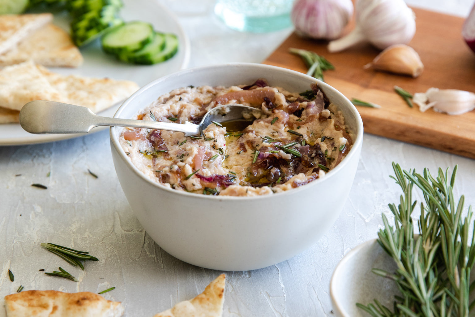 Bowl of caramelized onion white bean dip with spoon, garnished with rosemary and olive oil, served with crackers