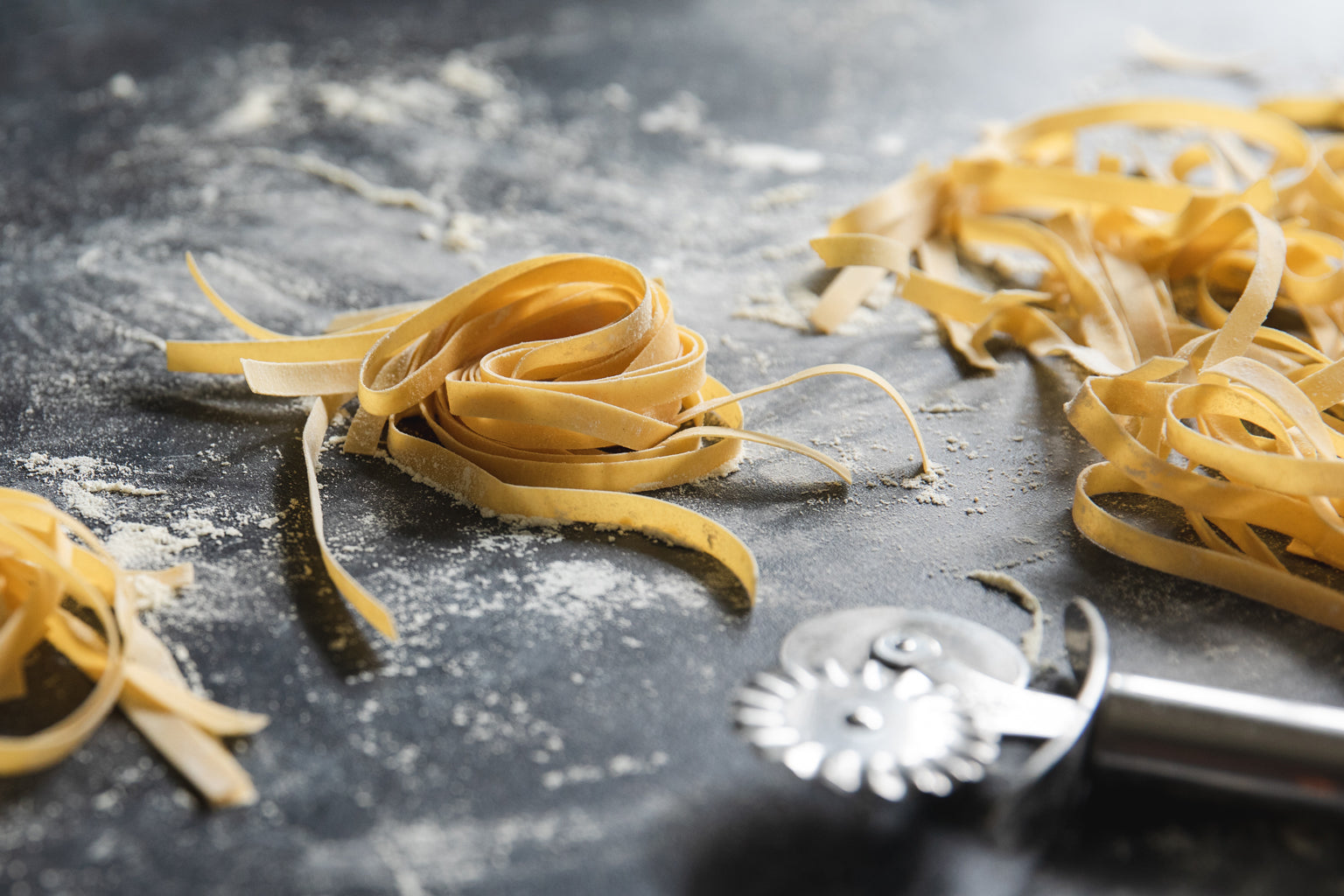Close-up of fresh Einkorn fettuccine nests showcasing hand-cut ribbon strands on a floured dark surface with a pasta cutter