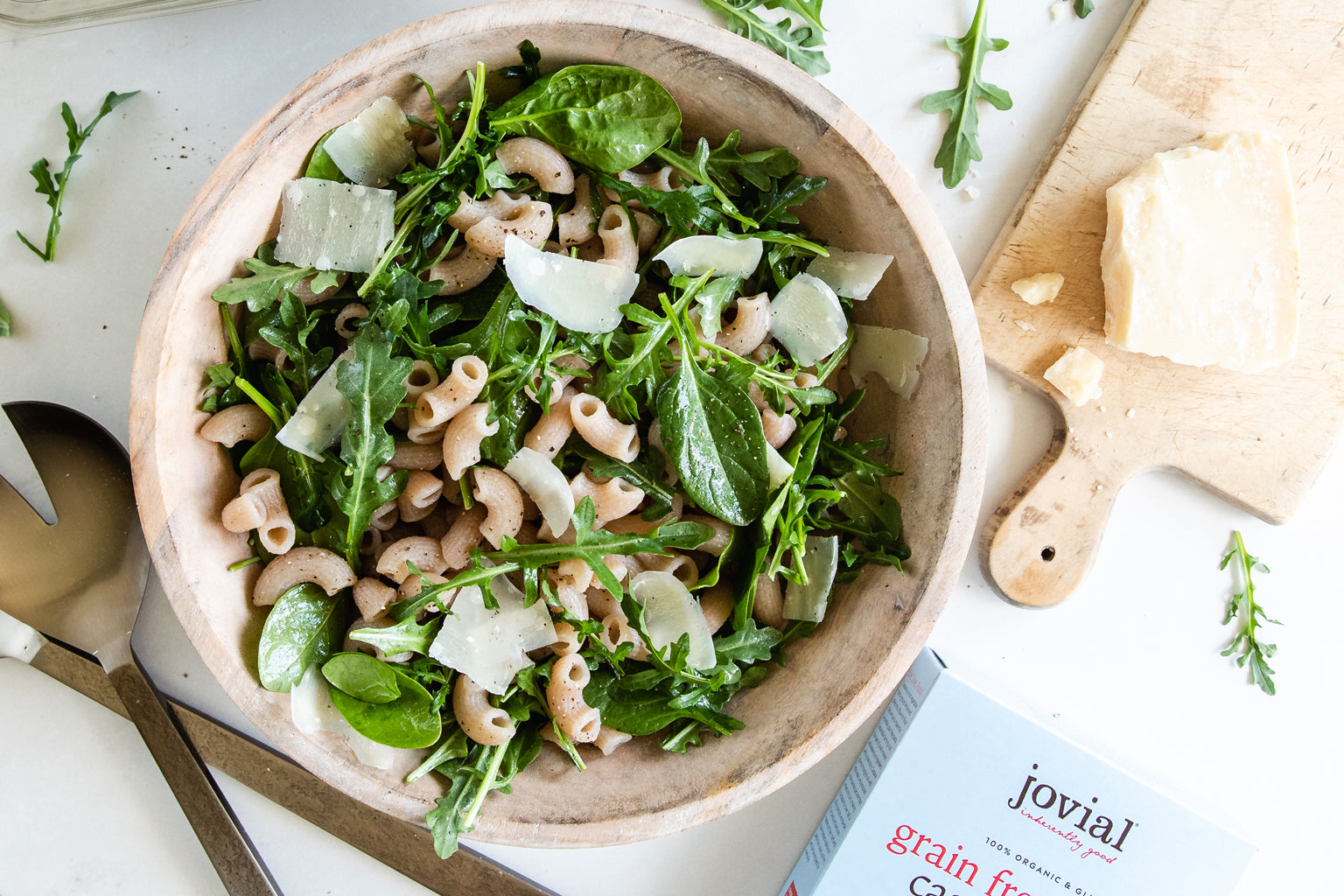 Pasta salad tossed with cassava elbows, baby spinach and arugula, and shaved Parmigiano in a wooden bowl on a white counter