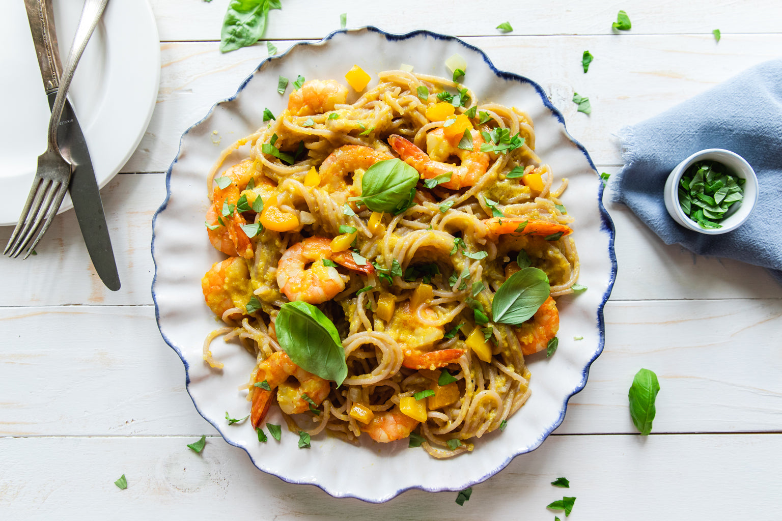 Cassava noodles with shrimp and yellow pepper plated on a scalloped white plate, garnished with basil on a light wooden table