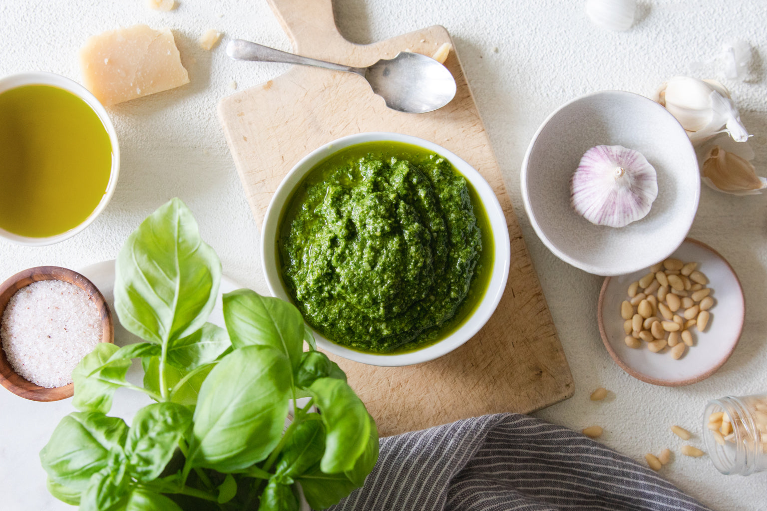 Bowl of basil pesto on a wooden cutting board with basil sprigs, pine nuts, garlic, Parmesan, olive oil, and cloth