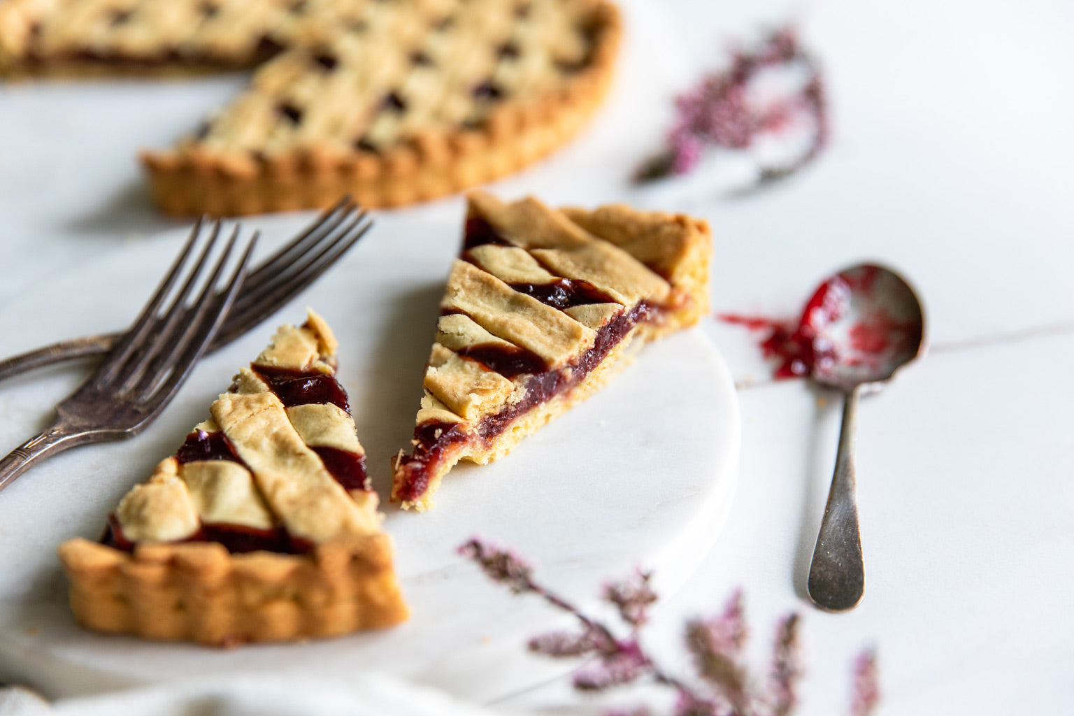 Two slices of Einkorn sour cherry crostata with lattice crust on a marble serving board, forks and a jam-covered spoon in soft natural light