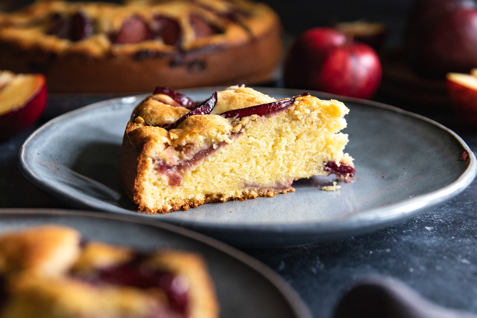 Side view of a slice of einkorn plum cake showing moist crumb and plum slices on a gray plate with blurred plums in back